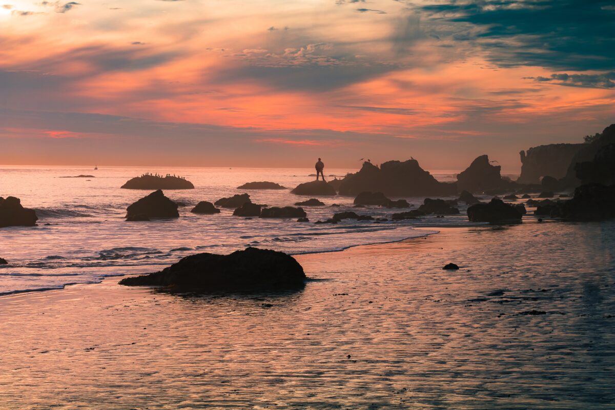 Sunset picture of Malibu beach with a person standing in the distance on a rock in the ocean. Pink and orange sunset clouds.