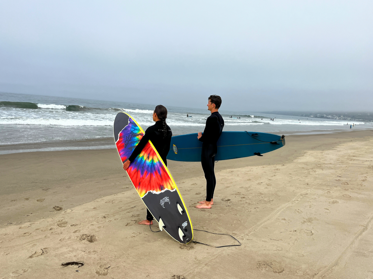 Two surfers at Zuma Beach, a girl and guy, looking out at the ocean holding their boards. One board is tie dye. 