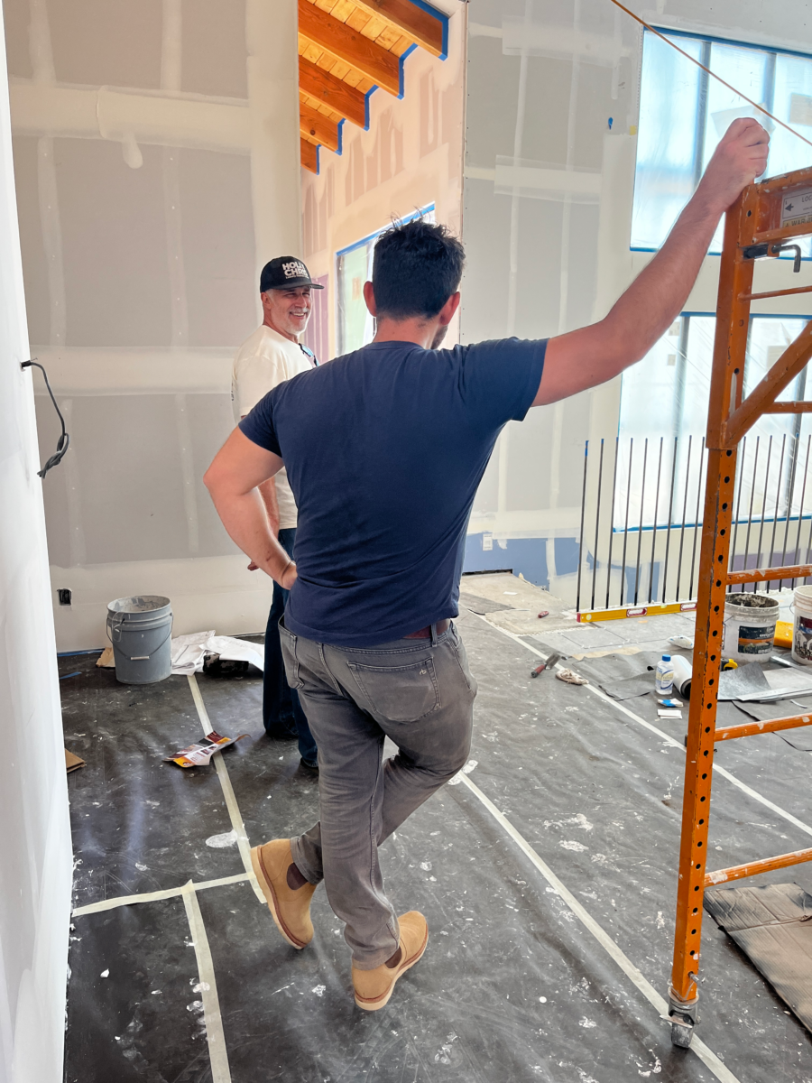 One man with his hand on his hip and the other holding onto scaffolding with his legs crossed talking to another man in the background laughing, in what appears to be a construction site.